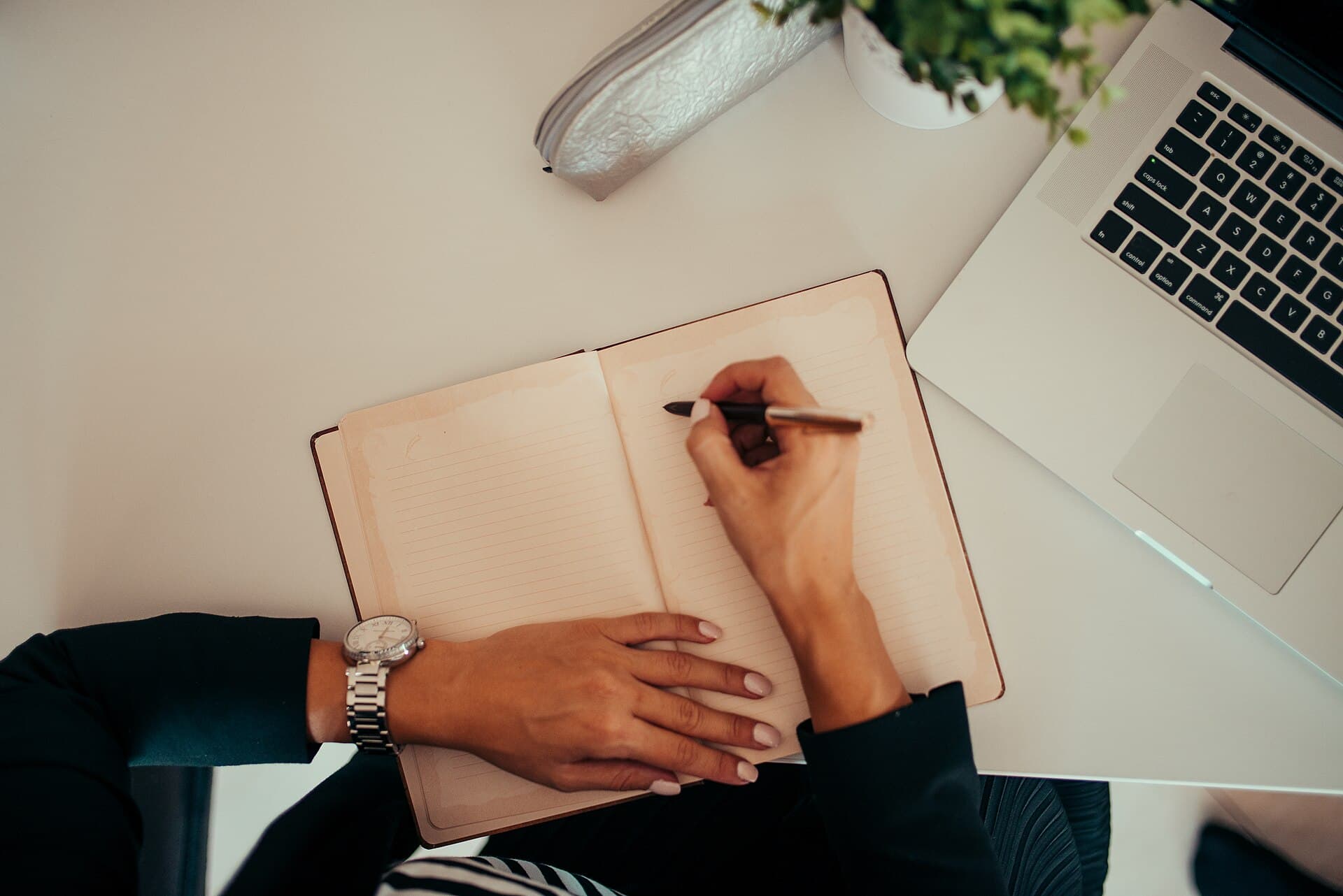 A person writing in a notebook beside a laptop — drafting an IELTS Writing Task 2 essay